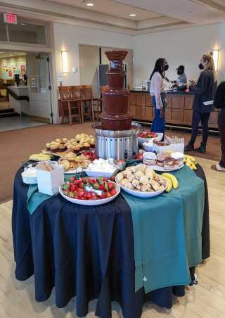 Im Vordergrund steht ein Tisch mit einem dunkelblauen Tischtuch. Auf dem Tisch steht ein grosser Schokoladenbrunnen, umgeben von einer Auswahl an Obst, Muffins und Gebäck. Im Hintergrund stehen Menschen an einem Buffet.