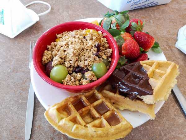 Ein Teller mit Essen auf dem Tisch: frische Waffeln mit Schokolade, frische Erdbeeren und eine Schale mit Trauben und Granola.