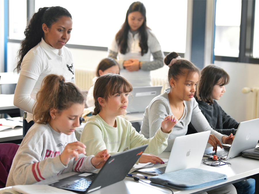 Mädchen arbeiten von vorne gesehen an einem Laptop und eine studentische Hilfskraft steht hinter ihnen