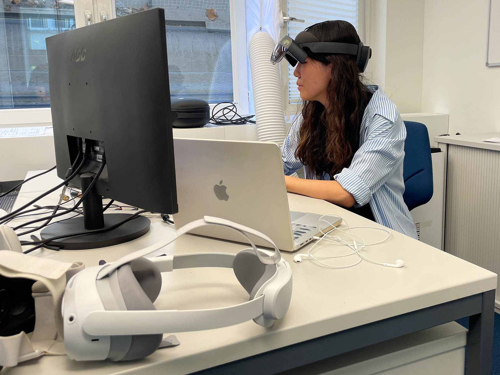 Portia sitzt an ihrem Schreibtisch und trägt eine AR-Brille. Im Vordergrund des Bildes ist eine VR-Brille zu sehen.