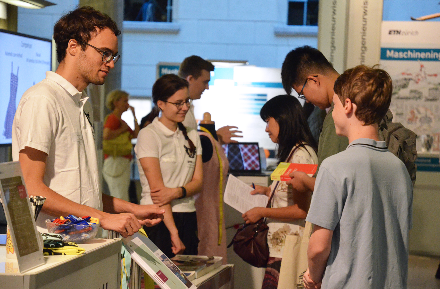 A student and a young pupil are standing opposite each other. The student is standing next to a bar table on which a bowl of chocolate is placed. In the background, other students are talking to visitors.