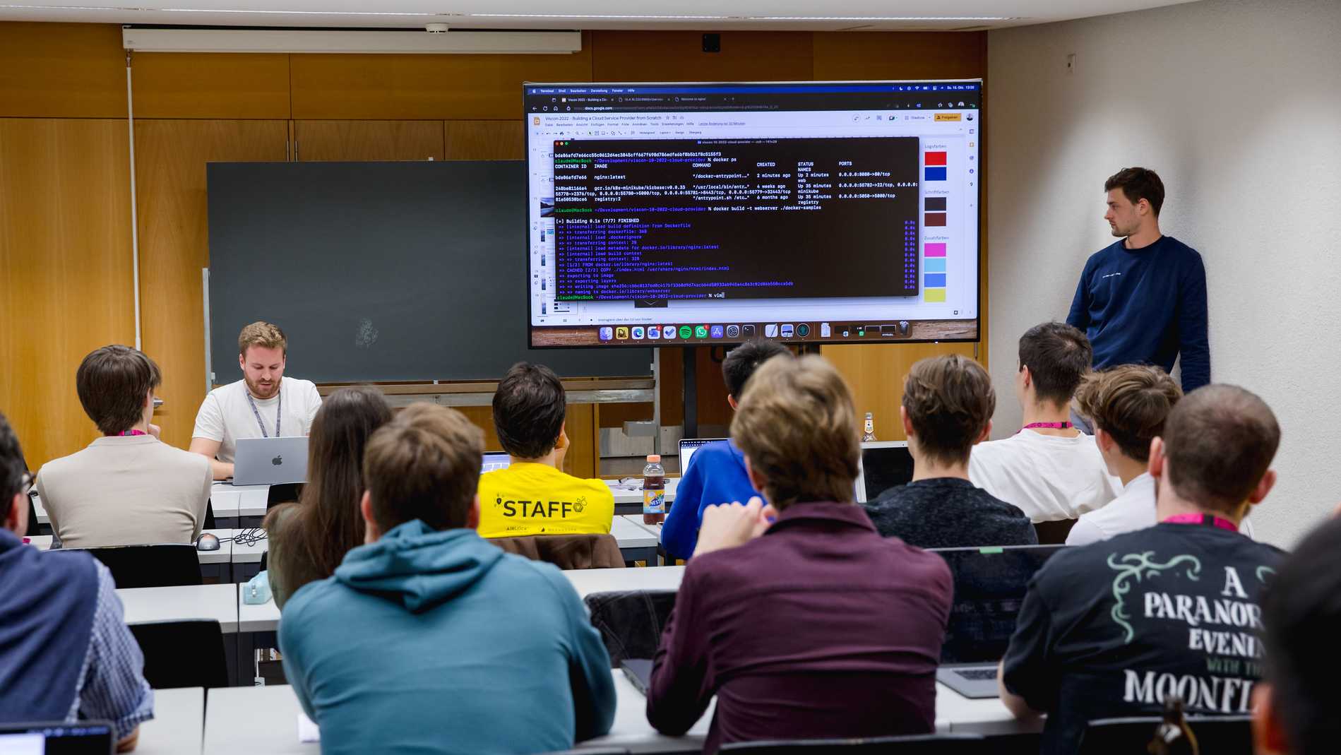 Students sit in a lecture hall and listen to two lecturers. There is a big screen in the front of the room where code is projected.