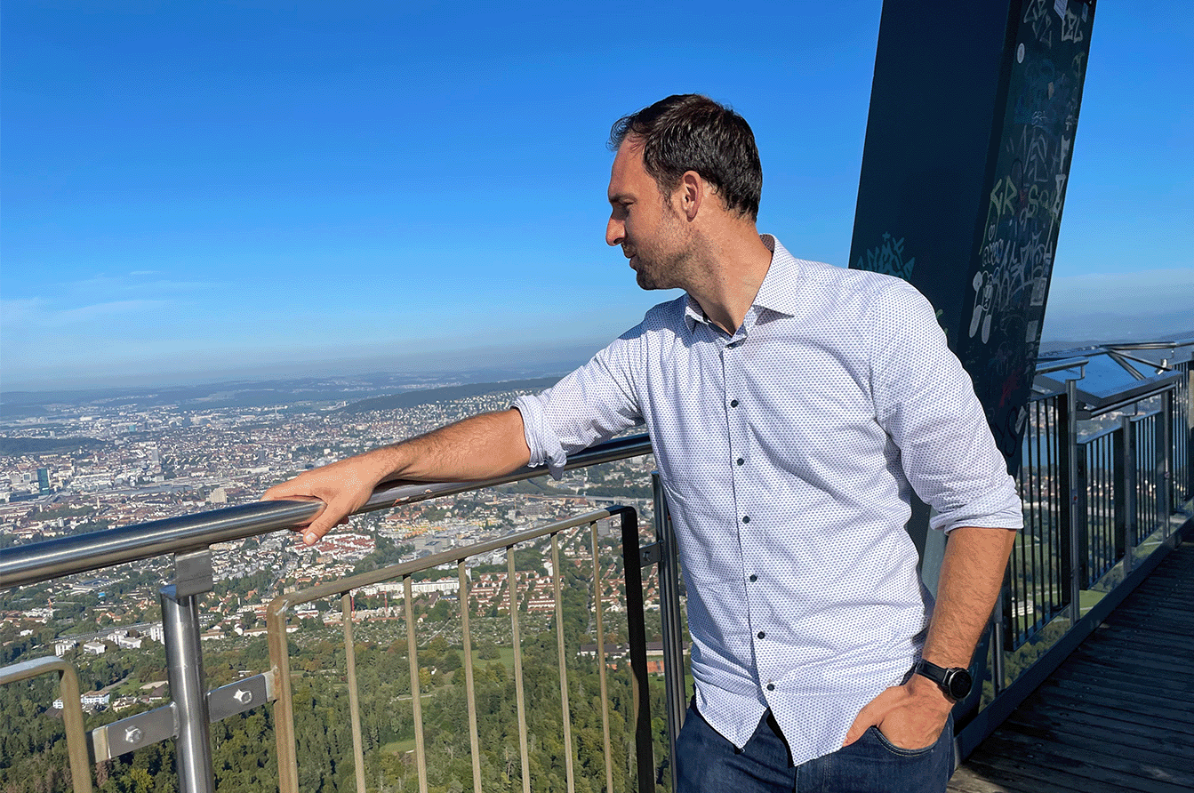 A man is standing on an observation platform. His hand is placed on the handrail and he looks to the left at the view of the city of Zurich.