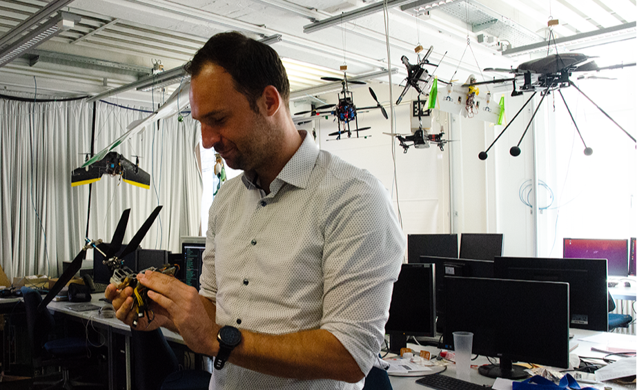 A man stands in the foreground and looks at a drone in his hands. More drones are hanging from the ceiling, attached with ropes. Tables with computers can be seen in the background.